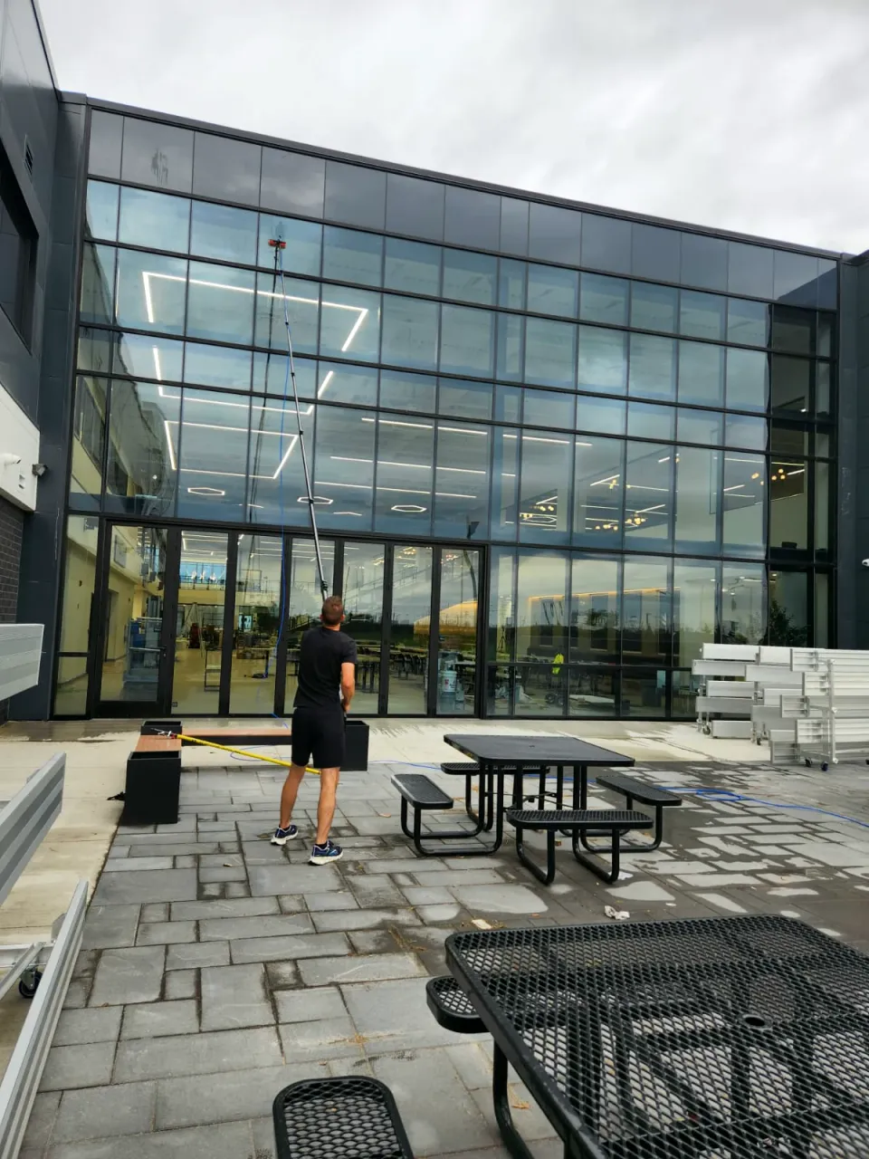 Window cleaner using a water-fed pole on a two-storey glass wall in a commercial courtyard with picnic tables