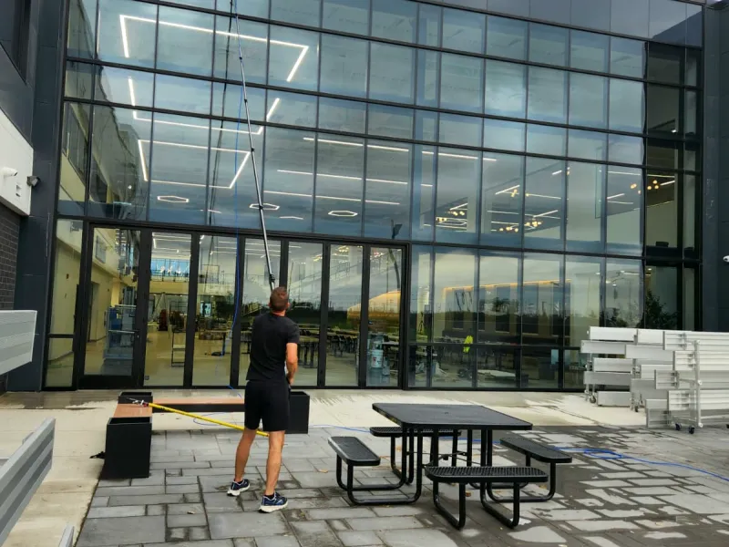 Window cleaner using a water-fed pole on a two-storey glass wall in a commercial courtyard with picnic tables