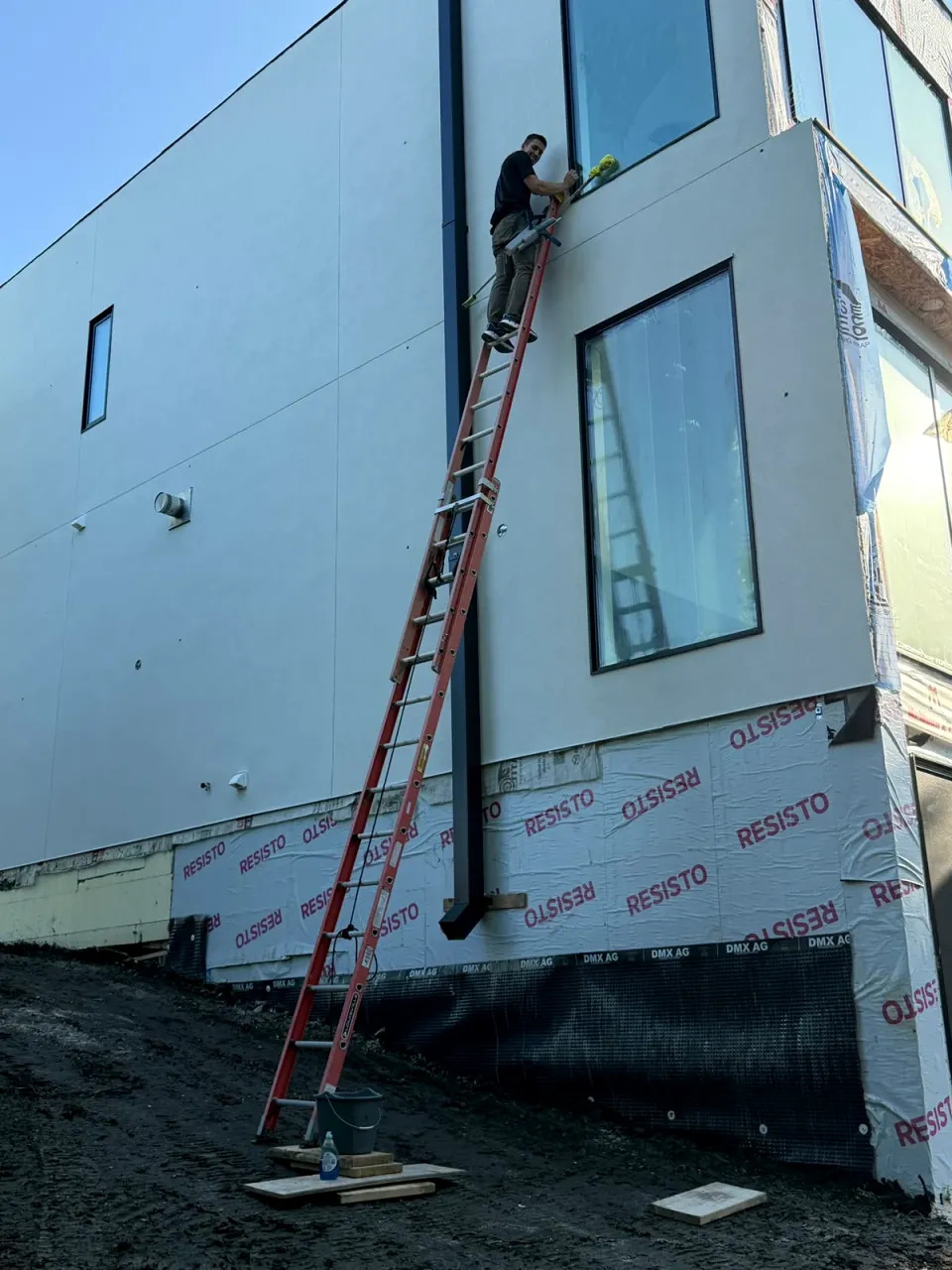 Window cleaner on a tall ladder cleaning an upper window on the outside of a new building still in house wrap