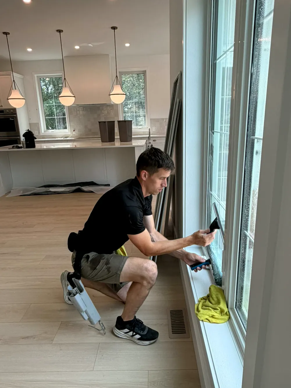 Marko kneeling at a tall window with a squeegee and scrubber in a bright new kitchen with protective sheeting on the floor
