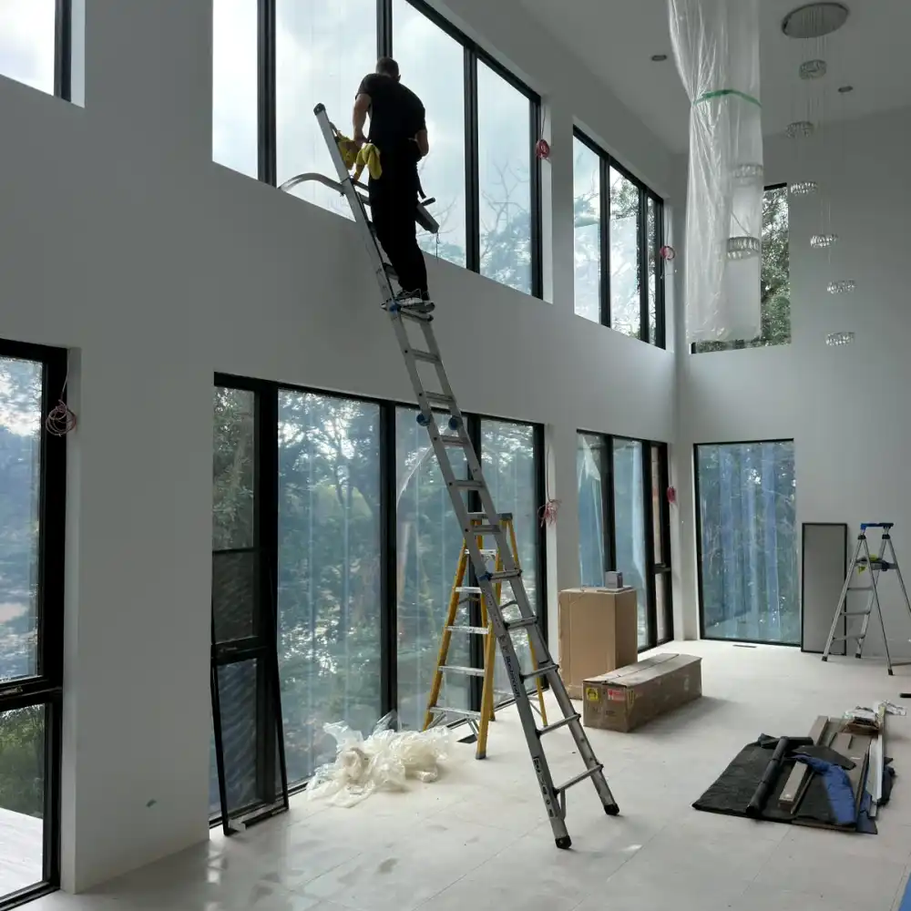 Window cleaner on a ladder cleaning high windows in a newly built room, with packing boxes and plastic on the floor