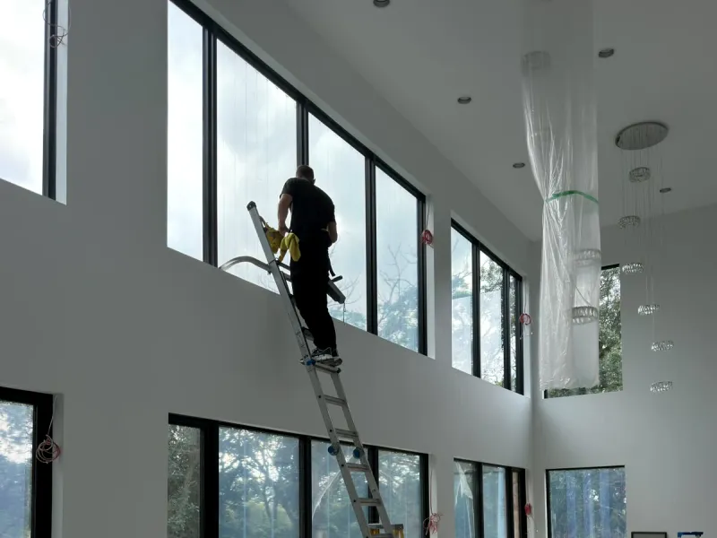 Window cleaner on a ladder cleaning high windows in a newly built room, with packing boxes and plastic on the floor