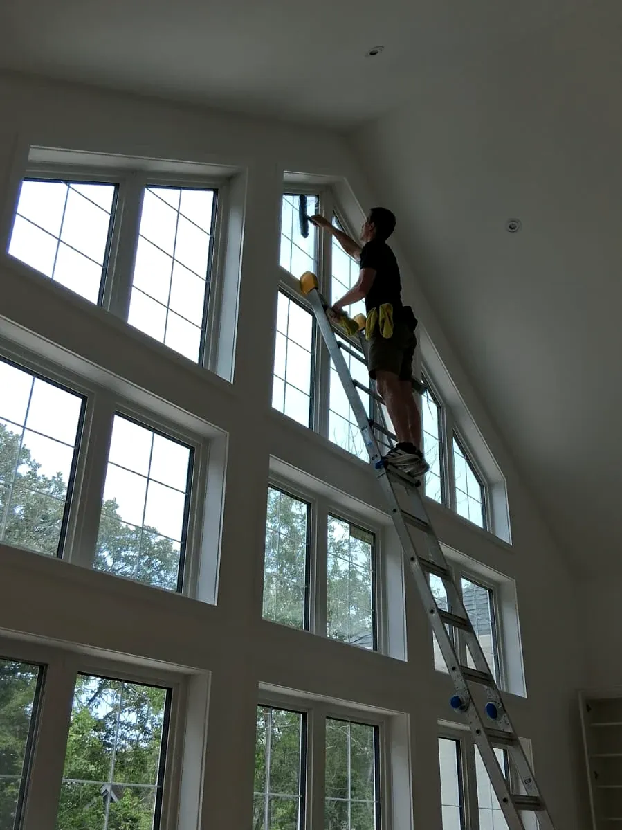Window cleaner on a ladder squeegeeing the top row of windows in a room with a tall vaulted wall of glass